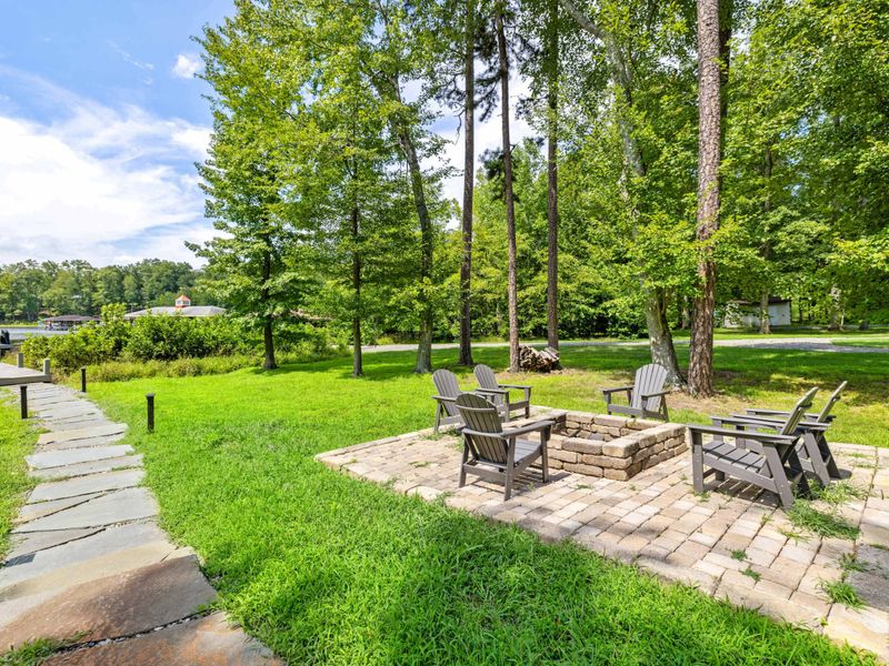 A cozy outdoor fire pit area surrounded by Adirondack chairs, set on a stone patio with lush greenery and easy access to the lake.