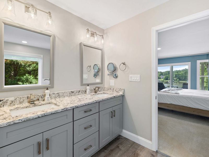 A bright and modern bathroom with a dual-sink vanity, granite countertops, and a view leading to the serene bedroom with lake vistas.