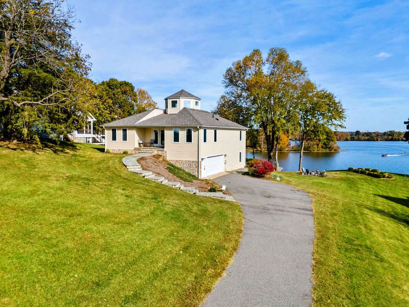 Aerial - Drone view of the lakefront home with a cupola, stone foundation, winding driveway, and the lake stretching beyond.