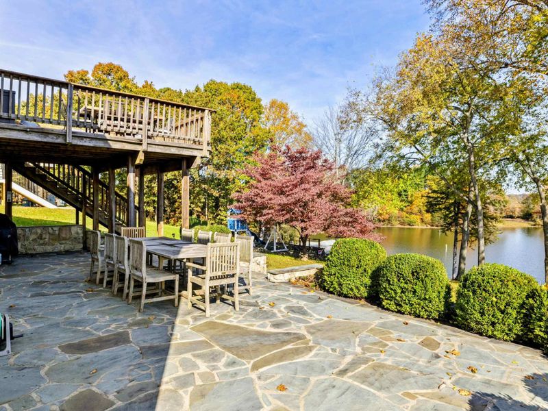 Expansive stone patio with dining area, surrounded by lush greenery and overlooking a serene lake