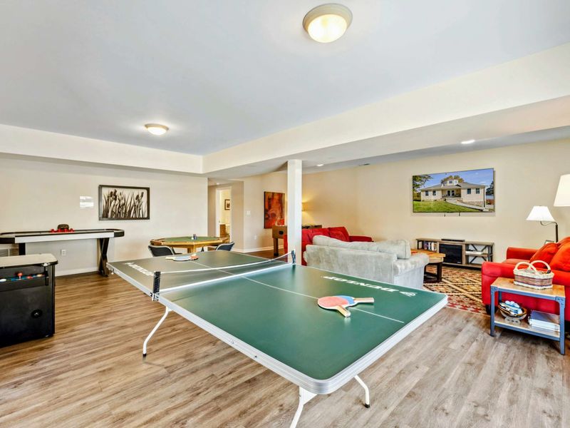 Game Room - Wide view of the lower level with ping pong, air hockey, bumper pool, red sofas, and a lounge area with wall-mounted TV.