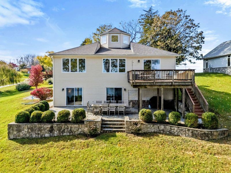 Elegant lakefront home with a large deck, stone patio, and manicured landscaping, ideal for enjoying outdoor spaces