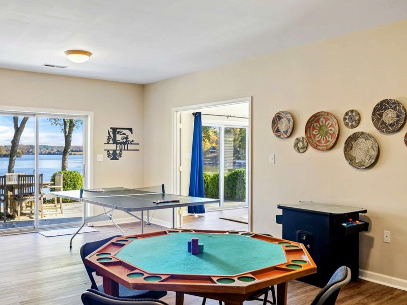 Game Room - Poker table, ping pong, and a retro arcade cabinet with woven basket wall art and lake views through the sliding doors.