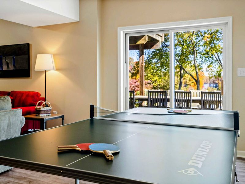 Game Room - Ping pong table with paddles ready for play, red sofa, and sliding glass doors to the autumn deck and lake beyond.
