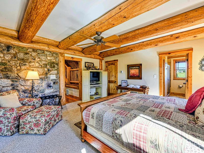 Relax in this spacious and cozy bedroom featuring rustic wood beams, a stone accent wall, comfortable seating, and charming decor for a perfect retreat.