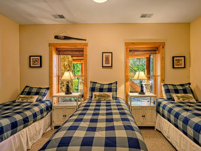 This charming bedroom offers three twin beds with cozy plaid bedding, warm wood accents, and natural light from two scenic windows.