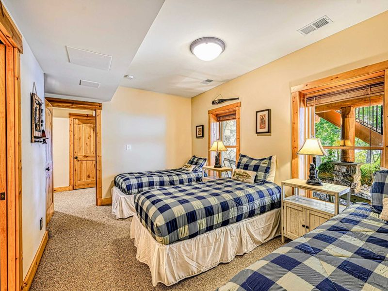 This charming bedroom offers three twin beds with cozy plaid bedding, warm wood accents, and natural light from two scenic windows.