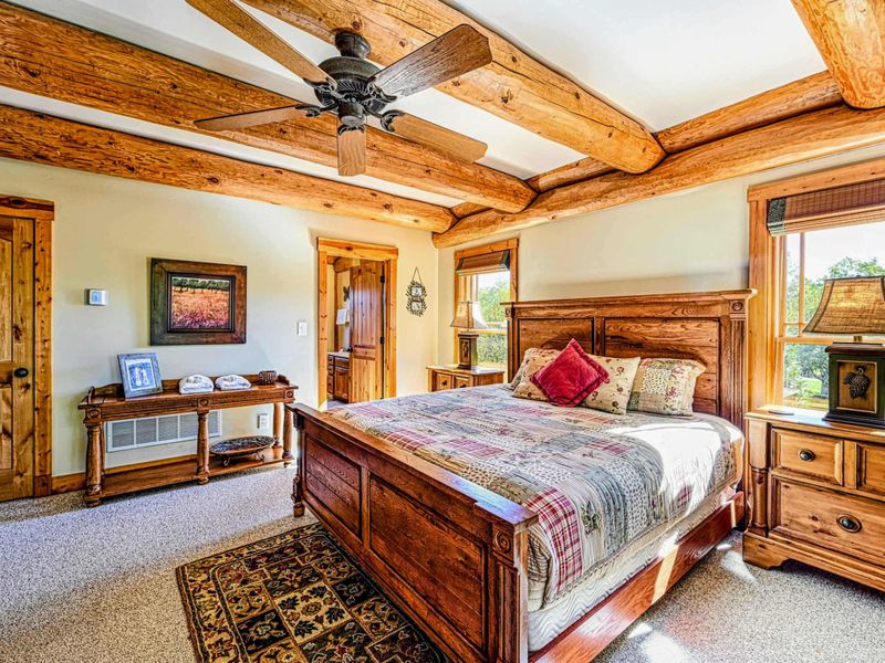Relax in this spacious and cozy bedroom featuring rustic wood beams, a stone accent wall, comfortable seating, and charming decor for a perfect retreat.