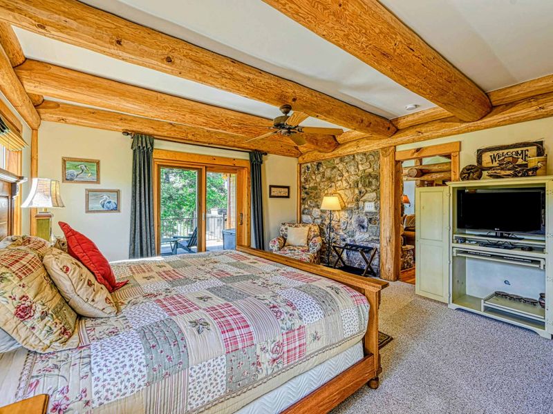 Relax in this spacious and cozy bedroom featuring rustic wood beams, a stone accent wall, comfortable seating, and charming decor for a perfect retreat.