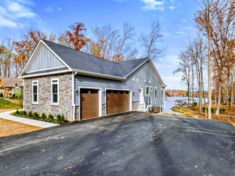 Spacious modern home with a stone and siding exterior, attached garage, and a long paved driveway leading to a peaceful wooded setting near the water.