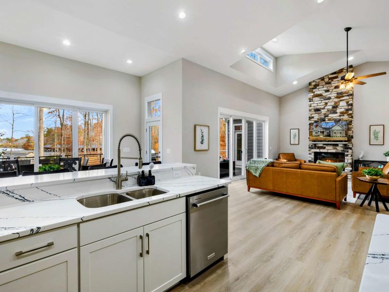 Bright open-concept kitchen with marble countertops and a view of the living area featuring a stone fireplace and large windows overlooking the outdoors.