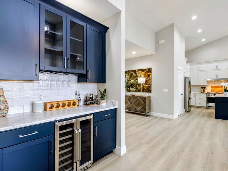 Modern wet bar with navy cabinets, a wine fridge, and a sleek countertop, seamlessly connected to a bright open kitchen and living area.