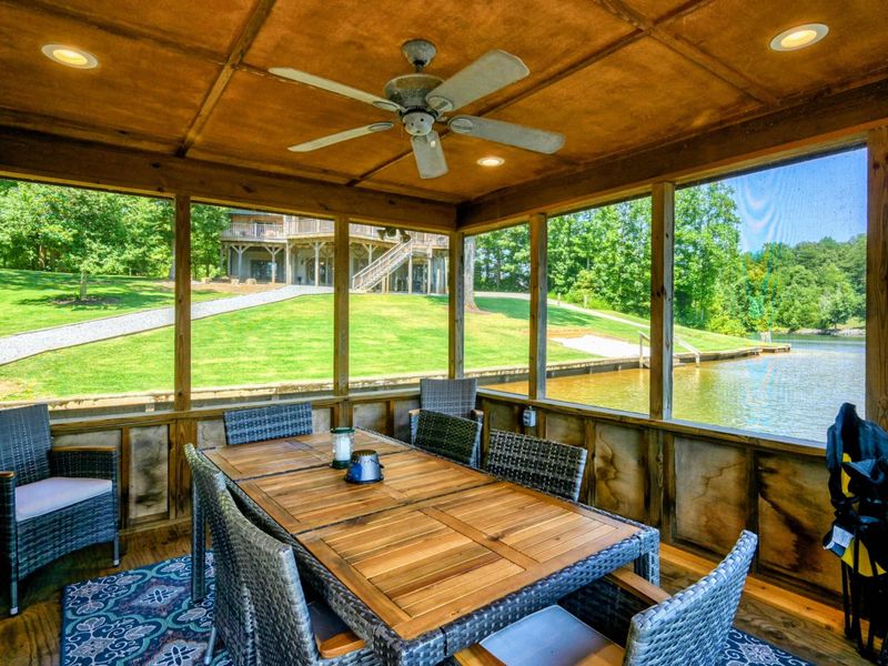 Screened-in area in the boathouse featuring a ceiling fan, comfortable seating, and a dining area with views of the lush green surroundings.