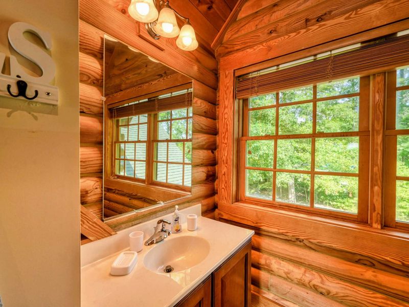 A cozy en suite bathroom features a walk-in shower, a single vanity with a large mirror, and rustic log cabin details complemented by natural light.