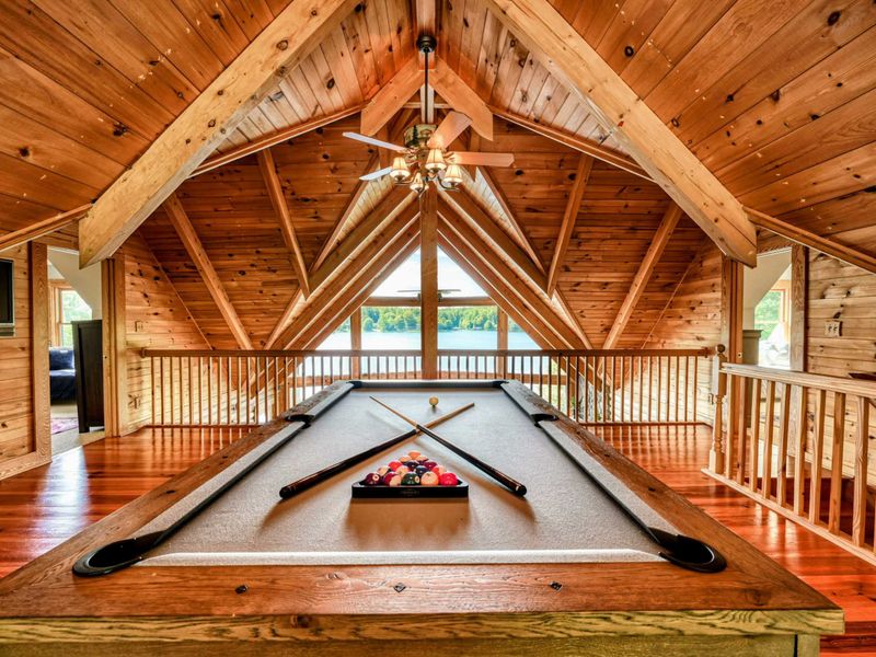 A beautifully crafted loft space features a pool table with lake views framed by vaulted wooden ceilings.