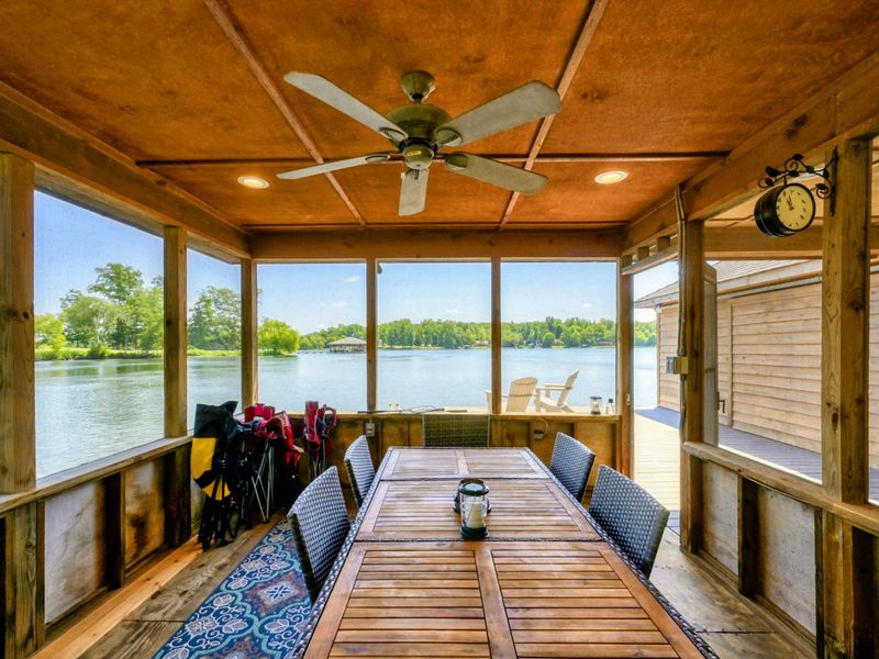 Screened-in area in the boathouse featuring a ceiling fan, comfortable seating, and a dining area with views of the lush green surroundings.