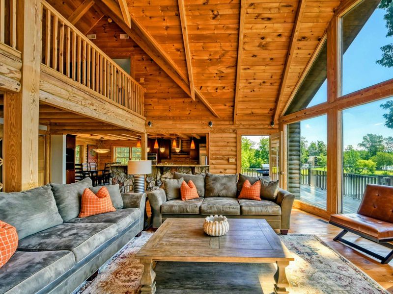 A warm and inviting open-concept living space features plush seating, vaulted wooden ceilings, and large windows with stunning lake views.