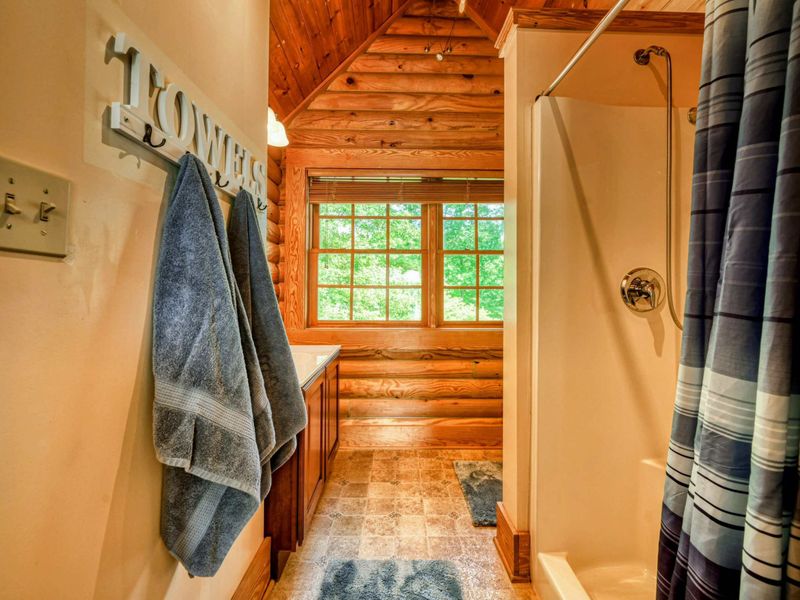 A cozy en suite bathroom features a walk-in shower, a single vanity with a large mirror, and rustic log cabin details complemented by natural light.