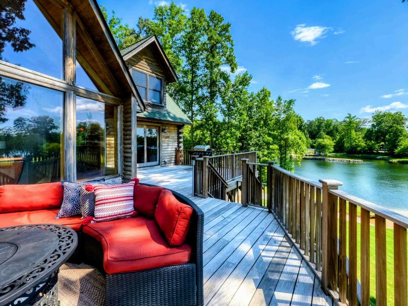 A stunning lakefront cabin features a spacious wooden deck with ample seating, surrounded by lush greenery and natural beauty.