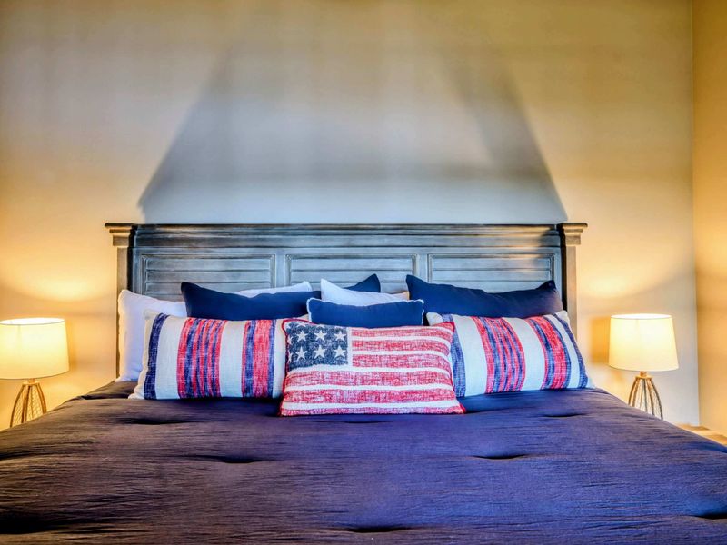 A stylish and cozy bedroom features a king-size bed with patriotic-themed decor, warm lighting, and a ceiling fan for added comfort.