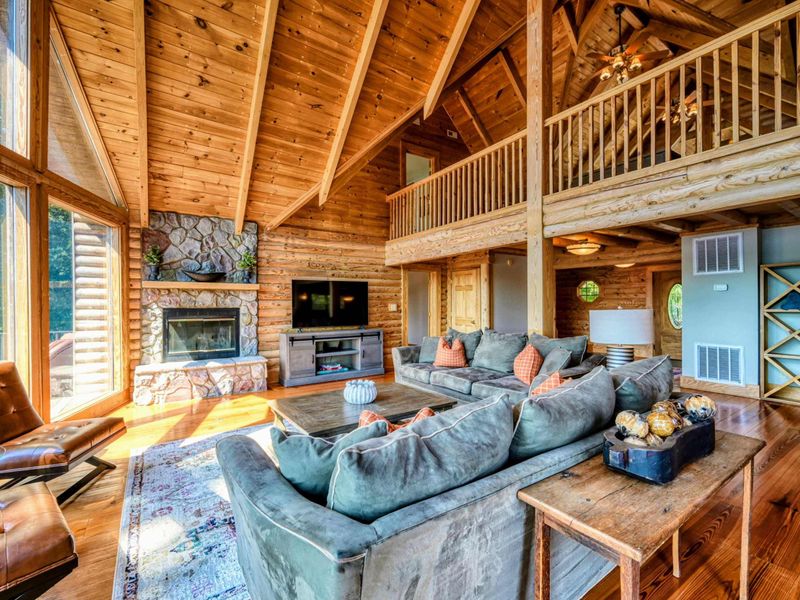 A warm and inviting open-concept living space features plush seating, vaulted wooden ceilings, and large windows with stunning lake views.