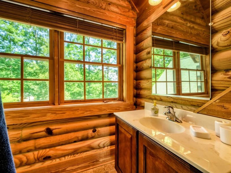 A cozy en suite bathroom features a walk-in shower, a spacious vanity with a large mirror, and charming log cabin accents with natural light streaming through the windows.