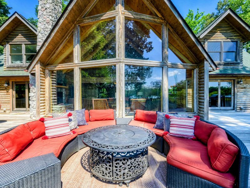 A beautifully designed outdoor seating area with vibrant cushions and a fire table, set against a rustic log cabin with expansive glass windows.