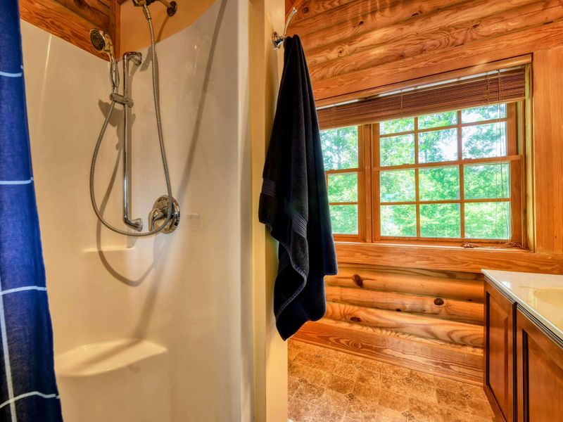 A cozy en suite bathroom features a walk-in shower, a spacious vanity with a large mirror, and charming log cabin accents with natural light streaming through the windows.
