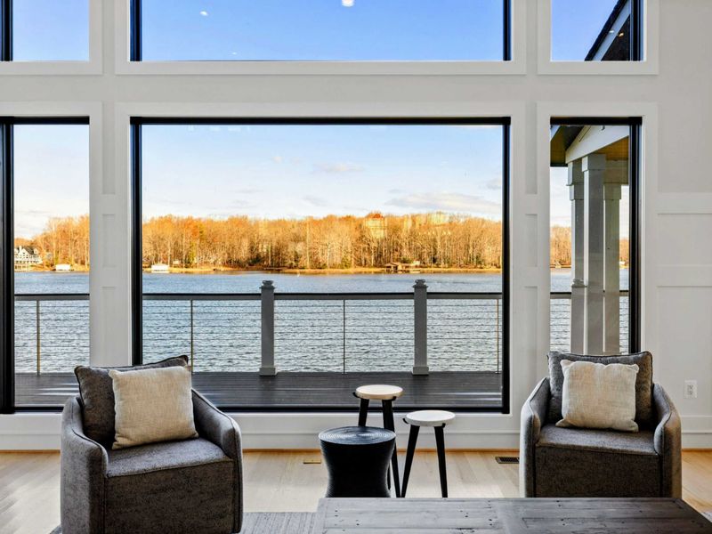 This stunning great room features soaring windows with breathtaking lake views, plush sectional seating, a modern fireplace, and seamless access to the kitchen and dining areas, creating the perfect space for relaxation and entertaining.