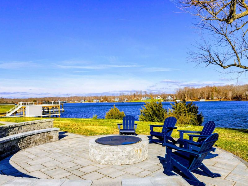 A scenic lakeside fire pit area with Adirondack chairs and a paved patio, offering a perfect spot to relax and enjoy the water views.