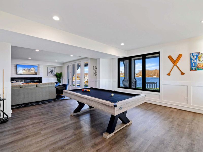 This spacious game room features a comfortable seating area with a modern fireplace, a pool table, ping-pong table, foosball table, shuffleboard, and a well-equipped kitchenette, all with stunning lake views.