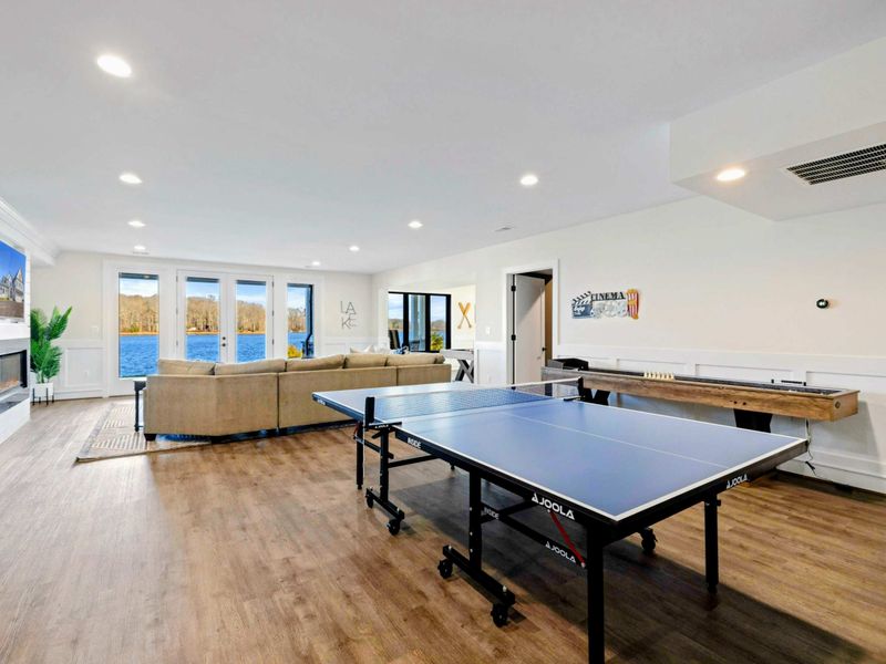 This spacious game room features a comfortable seating area with a modern fireplace, a pool table, ping-pong table, foosball table, shuffleboard, and a well-equipped kitchenette, all with stunning lake views.