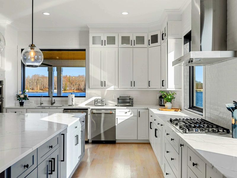 This modern and spacious kitchen features a large island with bar seating, elegant cabinetry, high-end appliances, and expansive windows showcasing scenic lake views, seamlessly flowing into the open concept living room.