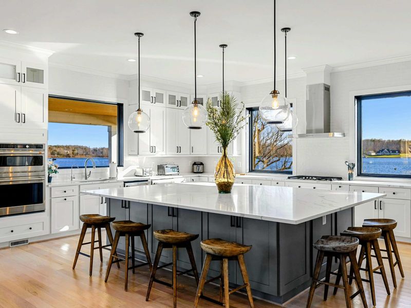This modern and spacious kitchen features a large island with bar seating, elegant cabinetry, high-end appliances, and expansive windows showcasing scenic lake views, seamlessly flowing into the open concept living room.