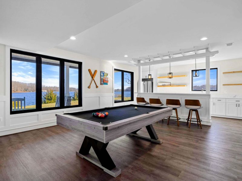 This spacious game room features a comfortable seating area with a modern fireplace, a pool table, ping-pong table, foosball table, shuffleboard, and a well-equipped kitchenette, all with stunning lake views.
