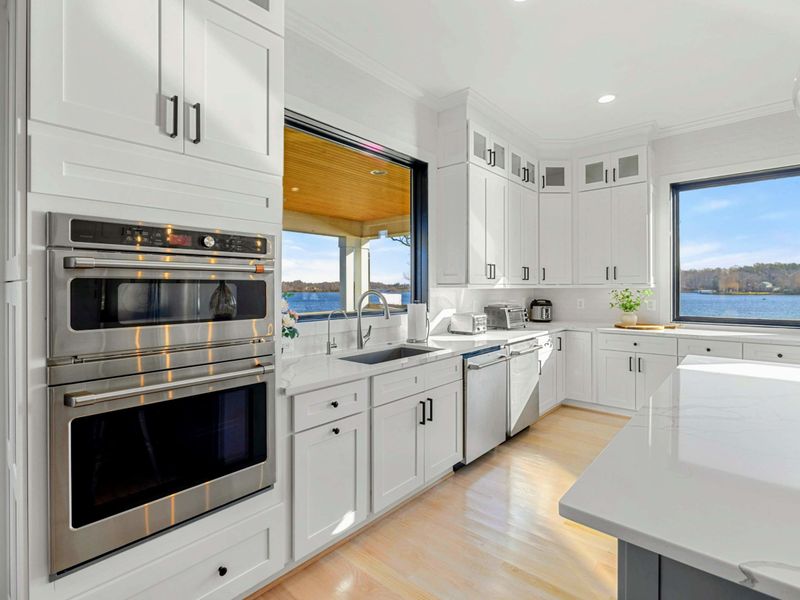 This modern and spacious kitchen features a large island with bar seating, elegant cabinetry, high-end appliances, and expansive windows showcasing scenic lake views, seamlessly flowing into the open concept living room.