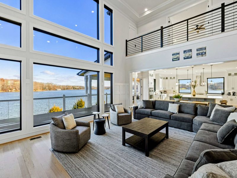 This stunning great room features soaring windows with breathtaking lake views, plush sectional seating, a modern fireplace, and seamless access to the kitchen and dining areas, creating the perfect space for relaxation and entertaining.