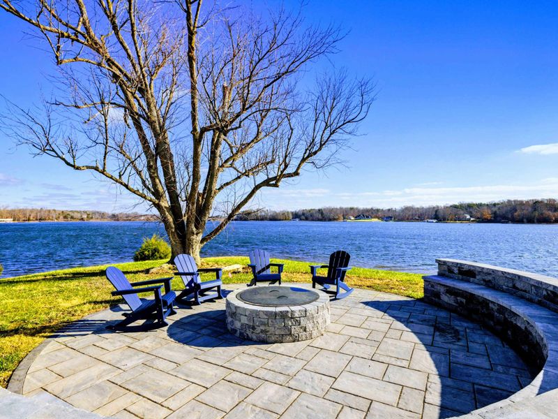 A scenic lakeside fire pit area with Adirondack chairs and a paved patio, offering a perfect spot to relax and enjoy the water views.