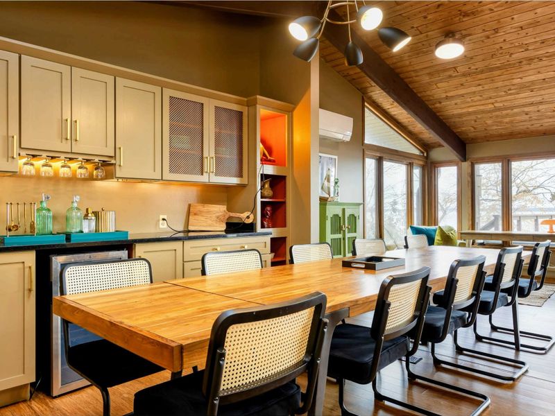 The dining area features a stylish wooden table that comfortably seats your entire group, complemented by modern cabinetry with built-in glass racks and a mini-bar setup, all while enjoying panoramic views through expansive windows.