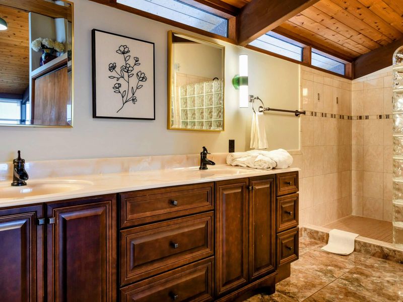 The primary suite bathroom features a double vanity with rich wood cabinetry, a large walk-in shower with glass block accents, and ample natural light, creating a serene and functional space.