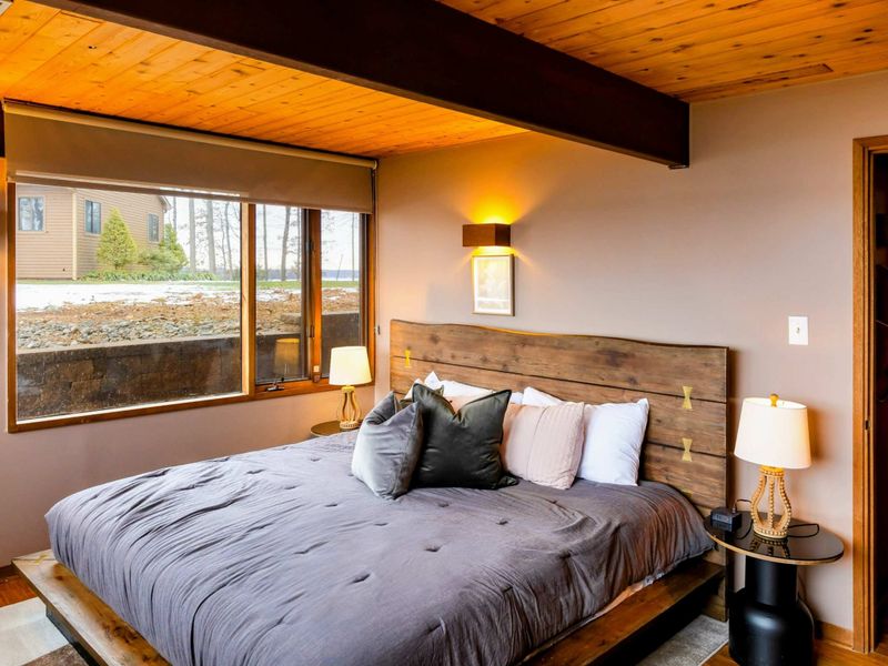 This bedroom features a rustic wooden bed frame, cozy bedding, and large windows inviting natural light and peaceful outdoor views.