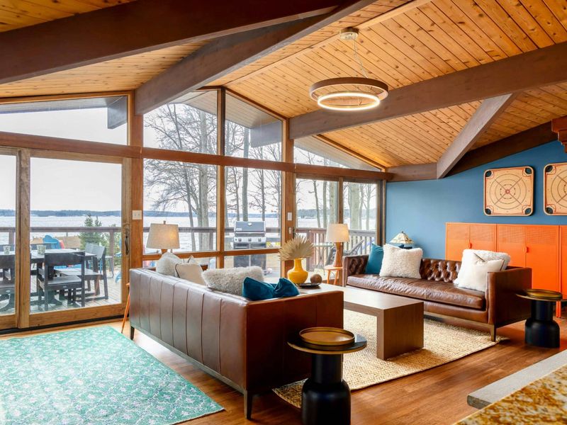 The Great Room offers a stylish retreat with cozy leather couches, vaulted wooden ceilings, and expansive glass doors that invite stunning lake views and natural light, seamlessly connecting the indoors with the outdoor deck.