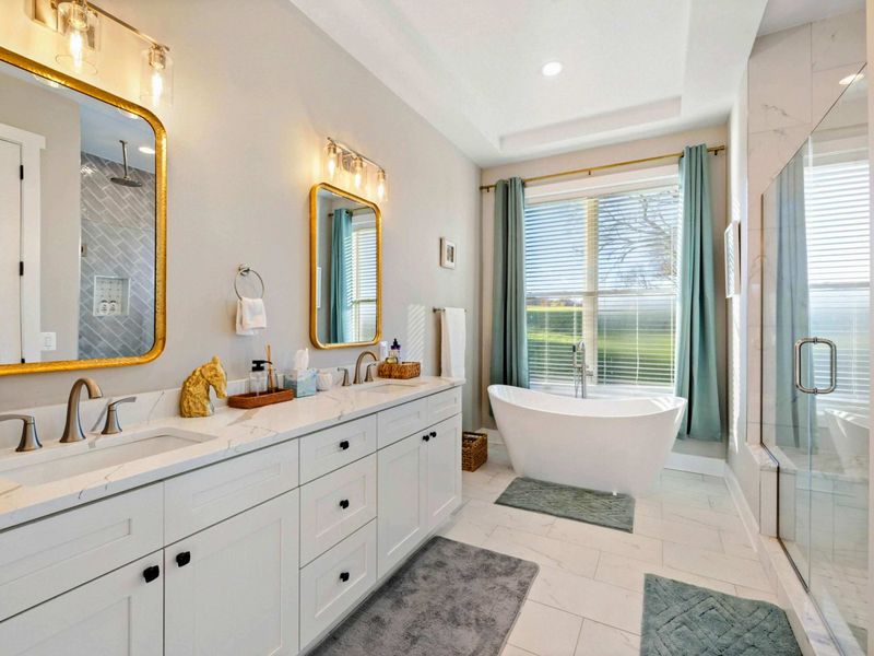 A spacious and elegant primary bathroom featuring a double vanity with gold-framed mirrors, a freestanding soaking tub, and a glass-enclosed walk-in shower with natural light.