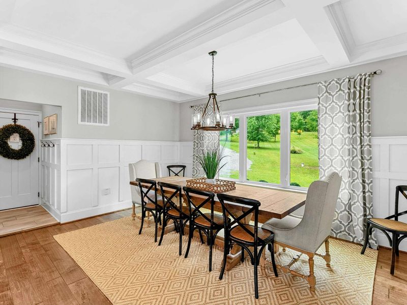 A bright and elegant dining room with a large farmhouse table, stylish seating, coffered ceilings, and a window offering scenic views of the backyard.
