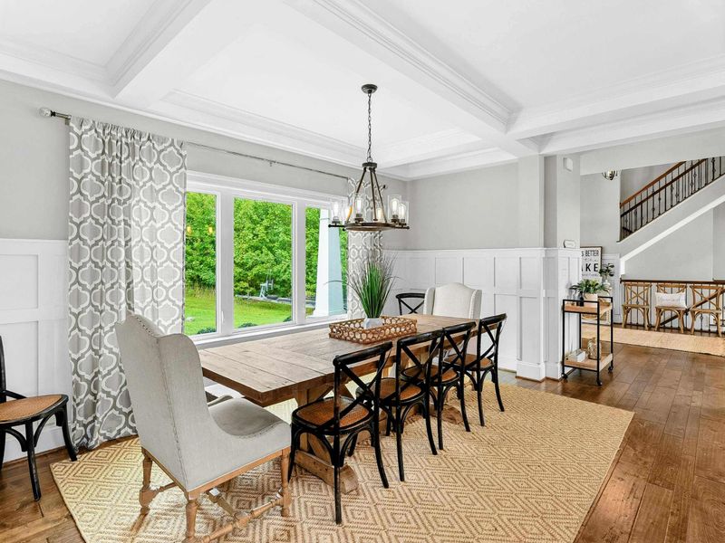 A bright and elegant dining room with a large farmhouse table, stylish seating, coffered ceilings, and a window offering scenic views of the backyard.