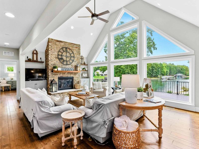A bright and spacious lakefront living area with large floor-to-ceiling windows, cozy seating, a stone fireplace, and open access to the kitchen and outdoor deck.