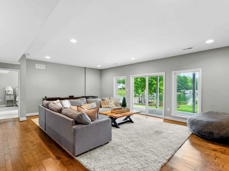 A spacious and inviting lower-level living area with a large sectional, rustic wood accent wall, entertainment center, and access to the backyard and lake views, complete with an adjacent bar and kitchenette.