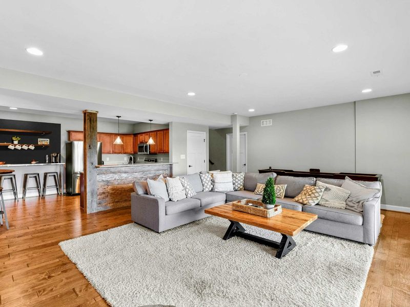 A spacious and inviting lower-level living area with a large sectional, rustic wood accent wall, entertainment center, and access to the backyard and lake views, complete with an adjacent bar and kitchenette.