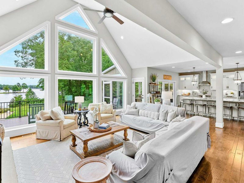 A bright and spacious lakefront living area with large floor-to-ceiling windows, cozy seating, a stone fireplace, and open access to the kitchen and outdoor deck.