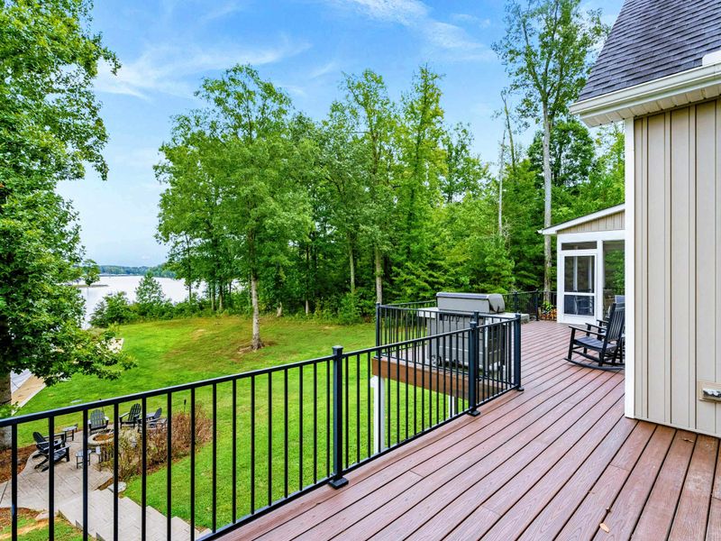A spacious outdoor deck with a serene lake view, ample seating, a grill for entertaining, and access to a screened-in porch and lush green yard.